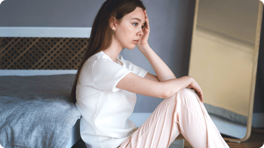 部屋でパジャマ姿の女性がベッドに背を持たれて悩む様子