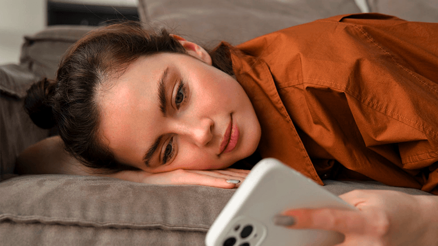ソファーに横たわりスマートフォンを見ながら考える女性