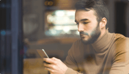 カフェの窓際でスマホを見つめる落ち着いた表情の男性。ベージュのタートルネックを着用
