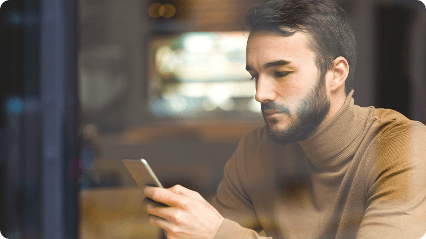 カフェの窓際でスマホを見つめる落ち着いた表情の男性。ベージュのタートルネックを着用