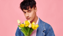 ピンクの背景で黄色いチューリップを手に持ち、優しく見つめる男性。恋の始まりを思わせる表情。