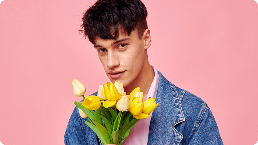 ピンクの背景で黄色いチューリップを手に持ち、優しく見つめる男性。恋の始まりを思わせる表情。