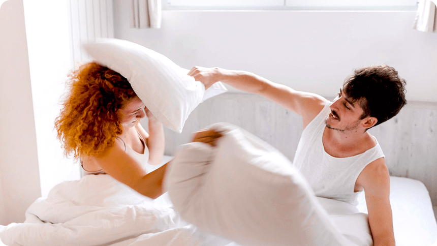 カップルがベッドの上で、楽しそうに枕をぶつけ合う姿