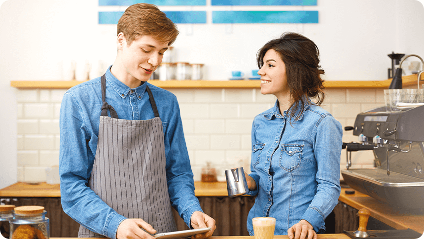 カフェで働く男女スタッフが笑顔で会話する様子。バイト先での自然なコミュニケーションを描いたシーン。