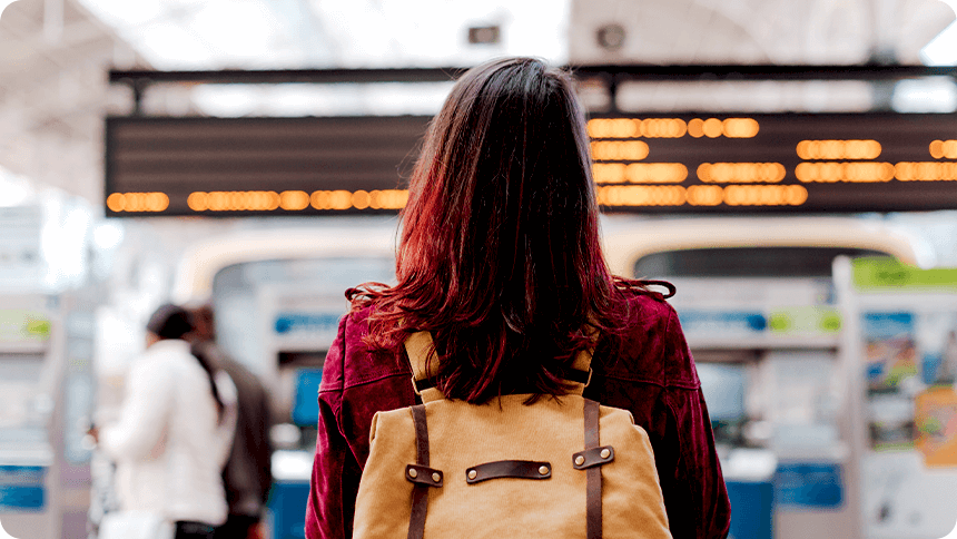 バックパックを背負い、改札前で電光掲示板を見上げる女性の後ろ姿。新しい出会いや旅立ちを感じさせる場面。