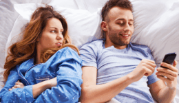 ベッドでスマホに夢中な男性を見て、不満そうに横目で見つめる女性。カップルのすれ違いを示す場面