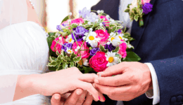 結婚式で新郎新婦が指輪を交換する瞬間の手元。華やかなブーケが背景に映る。