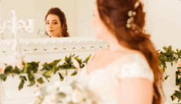 鏡の前に立つウェディングドレス姿の女性が、自分自身を静かに見つめている様子。白を基調とした室内には装飾用の花やグリーンが添えられている。