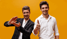 オレンジ背景の前で、スーツ姿の男性2人が並び、片方は拍手、もう片方は親指を立てている写真