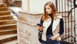 屋外の建物の前で、スマートフォンを見つめながら少し沈んだ表情を浮かべている女性。