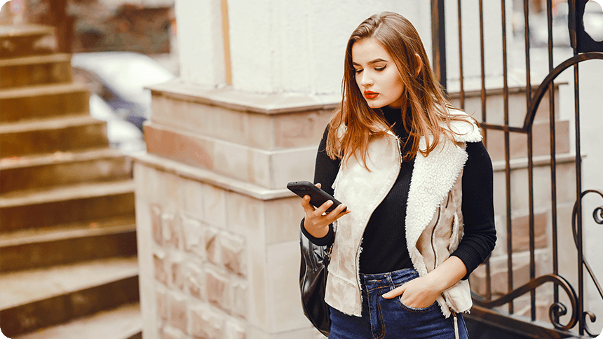 屋外の建物の前で、スマートフォンを見つめながら少し沈んだ表情を浮かべている女性。