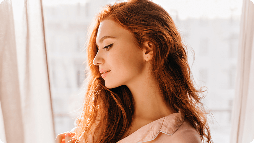 窓辺で目を閉じ、静かに微笑む女性。感受性を大切にしながら、無神経な人に振り回されずに生きる方法を見つめ直しているような表情。