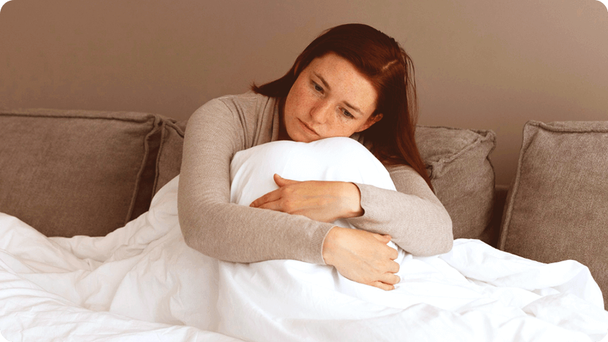 ベッドで膝にかかった布団ごと膝を抱える女性