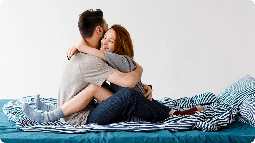 ベッドの上で幸せそうな笑みな彼女が、彼に抱えられるようにハグされているカップル。