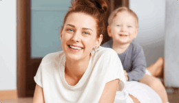 笑顔で床にうつ伏せになる女性と、その背中に乗って楽しそうに笑う幼い子ども。穏やかな室内で寄り添う親子の姿。