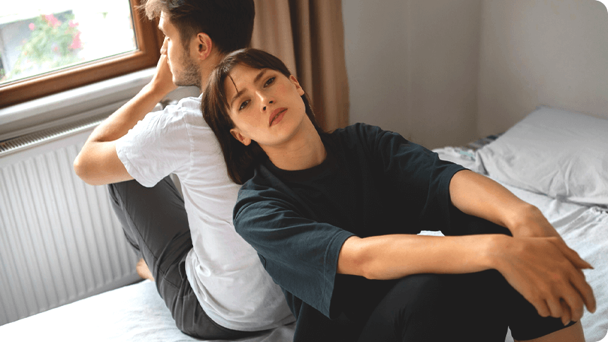 男性と背中合わせでベッドに座る女性の表情が少し曇っている