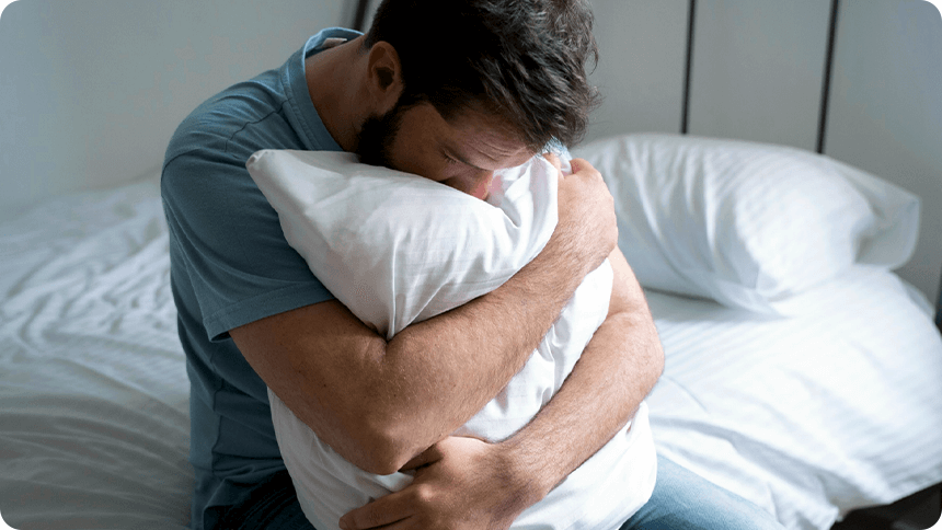 ベッドに腰を掛けながら枕を抱きしめ悩んでいる様子の男性