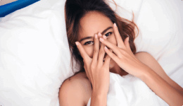 ベッドで布団をかぶり、手で顔を隠して笑う女性