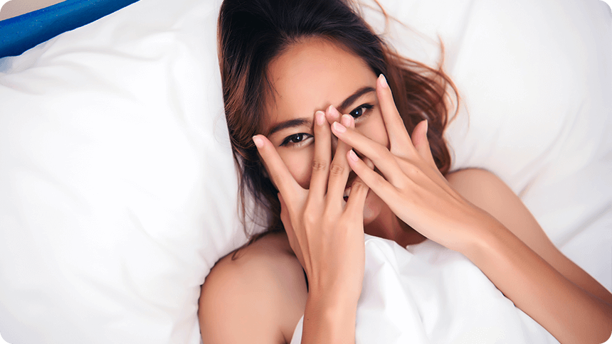 ベッドで布団をかぶり、手で顔を隠して笑う女性